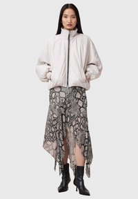 Lichtbeige bomberjack met een hoge kraag en een rits aan de voorkant, gecombineerd met een grijze asymmetrische rok met slangenprint, gedragen met zwarte hoge hakken.