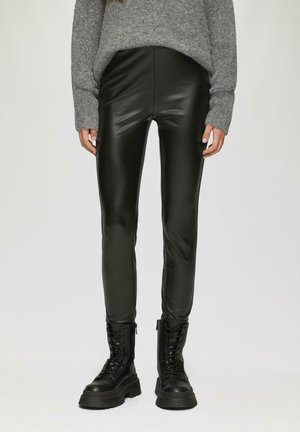 Zwarte nepleer leggings met een gladde glans, gecombineerd met een losse grijze gebreide trui en zwarte veterlaarzen.