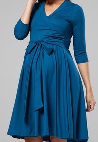 Tyrkisk omslagsklänning med V-ringning, långa ärmar och knytning i midjan. Tillverkad av mjukt, elastiskt tyg, har den en svängig kjol.