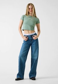 T-shirt vert court avec logo "GUESS" ; jean bleu taille haute à jambes larges ; chaussures noires ; design minimaliste sur un fond neutre.