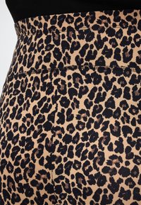 Ύφασμα με λεοπάρ τύπωμα σε εφαρμοστά, ψηλόμεσα κολάν ή παντελόνια με ορατές ραφές σε μπεζ φόντο.