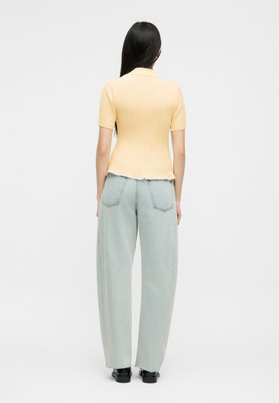 Γυναίκα με μακριά σκούρα μαλλιά, ντυμένη με ανοιχτό κίτρινο μπλουζάκι με κοντά μανίκια και ανοιχτό μπλε τζιν με φαρδιά παντελόνια, στέκεται γυρισμένη προς τα πίσω σε λευκό φόντο.