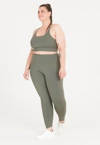Olivgrön träningsset bestående av en cropped sport-bh och hög midja leggings. Slät textur med sömdetaljer. Vita träningsskor.