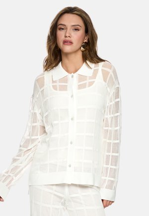 Woman wearing white button-up shirt with grid pattern mesh over matching pants, paired with large gold hoop earrings.