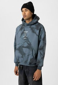 Pullover hoodie in donkerblauw-grijs tie-dye patroon, met een voorzak en witte opdruk. Gedirigeerd met zwarte jeans en een beanie.