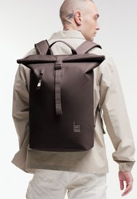 Hnědý batoh s rolovací sepnací klopou, nastavitelnými popruhy, minimalistickým designem, zipovou kapsou a logem v pravém dolním rohu.