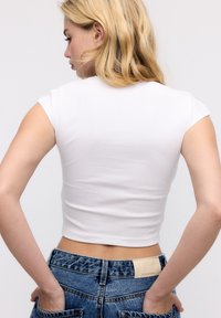 Femme blonde vue de dos, portant un crop top blanc ajusté et un jean bleu taille haute, les mains dans les poches arrière.