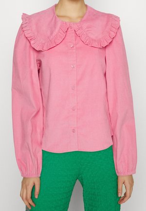 Femme portant un chemisier rose à manches longues avec un grand col à volants et un pantalon vert texturé, sur un fond uni.