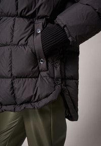 Chaqueta negra acolchada con botones a presión, mano en el bolsillo con puño de canalé, combinada con pantalones de cuero verde oliva.