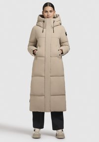 Garš beige pūkains mētelis ar pieguļošu kapuci, priekšējiem kabatām un melniem akcentiem. Gluda tekstūra ar horizontālām quilted sekcijām.