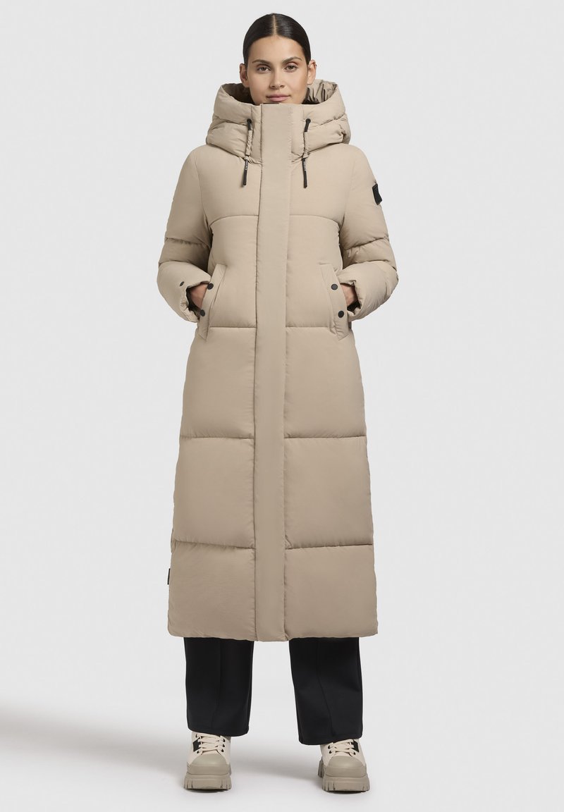 Garš beige pūkains mētelis ar pieguļošu kapuci, priekšējiem kabatām un melniem akcentiem. Gluda tekstūra ar horizontālām quilted sekcijām.