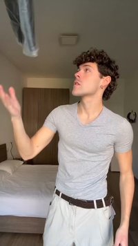 Tânăr îmbrăcat într-un tricou gri aruncând o pereche de blugi într-un dormitor cu un pat și un dulap de lemn în fundal.