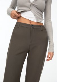 Brown tailored trousers with a smooth texture, featuring a front button closure and angled pockets, paired with a light gray ribbed cardigan.