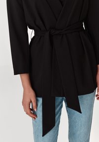 Svart överdel med lång ärm, knuten i midjan, ihop med ljusblå jeans. Tyget verkar mjukt med en slät textur.