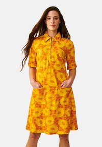 Keltainen mekko, jossa on oranssi kukkakuosi, lyhyet hihat, kaulus ja kaksi etutaskua. Vetoketjukiinnitys kaula-aukoissa.