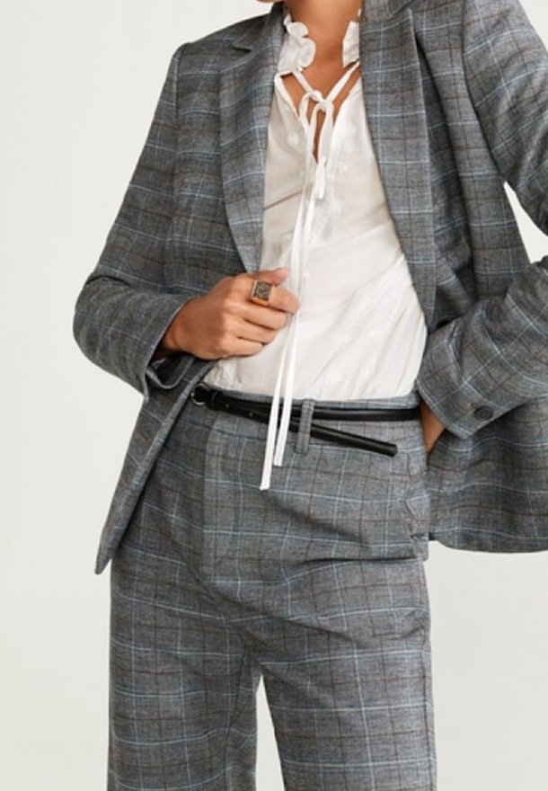 Costume à carreaux gris avec un blazer cintré et un pantalon assorti. Une blouse blanche en dessous avec un col lavallière. Une ceinture noire ajoute une touche de détail.
