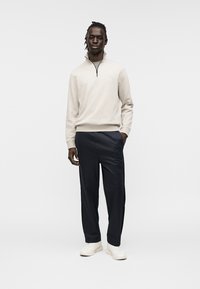 Beige Reißverschluss-Sweatshirt mit hohem Kragen, kombiniert mit dunkler Hose und weißen Sneakern. Glatter Stoff mit lockerem Sitz. Keine Muster oder Akzente.