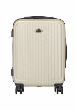 Beżowa walizka z twardą obudową z pionowymi żebrami, czarną plakietką z logo, cztery czarne obrotowe kółka oraz uchwyty po bokach i na górze.