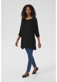 Tunique noire à coupe ample, avec un col froncé et des manches trois-quarts, associée à un jean skinny bleu et des ballerines noires.