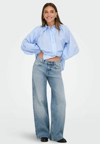 Camisa de botón azul claro, de corte sobredimensionado y con mangas voluminosas, combinada con pantalones vaqueros de tiro alto, de pierna ancha y azul deslavado, junto con botas negras de tobillo.