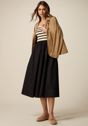 Frau trägt eine beige Jacke über einem schwarz-weiß gestreiften Top, einem schwarzen Midirock und beigen Mokassins, steht vor einem schlichten Hintergrund.