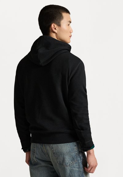 Svart bomull hettegenser med snorhette, ribbede mansjetter og en avslappet passform, kombinert med lyseblå denimjeans. Bakvisning.