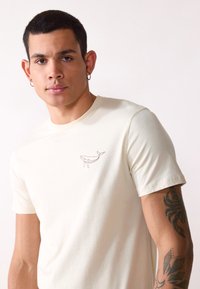 Light beige cotton t-shirt featuring a small brown whale embroidered on the left chest. Short sleeves, round neckline, and soft texture.