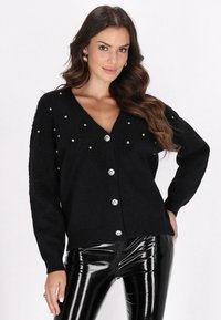 Cárdigan negro con escote en V y botones en la parte frontal, que presenta una tela texturizada y tachuelas plateadas dispersas. Llevado sobre pantalones negros brillantes.