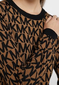 Jersey de punto con fondo marrón que presenta un patrón geométrico negro. Cuello y puños acanalados en negro. Textura suave.