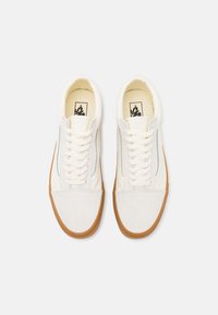 Białe sneakersy niskie z gumową podeszwą, białymi sznurowadłami i czarnym logo w środku na jednolitym jasnoszarym tle.