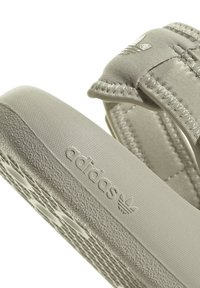 Svetlo sivi Adidas japonki s teksturiranim gumijastim podplatom, podstavljenim remenom i utisnutim logotipom sa strane. Mekana materijalna završna obrada.