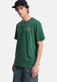 Jeune homme portant un t-shirt graphique vert foncé avec un motif de vague jaune, un short cargo gris foncé et une casquette noire, debout devant un fond blanc.