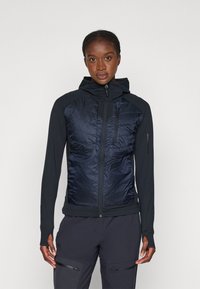Veste hybride avec un devant matelassé bleu marine et des manches noires extensibles. Dotée d'une poche poitrine zippée et d'une capuche ajustable pour un meilleur ajustement.