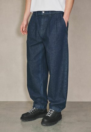 Person trägt lockere dunkelblaue Jeanshosen und schwarze Wanderstiefel mit weißen Schnürsenkeln, steht vor einem einfarbigen Hintergrund.