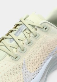 Αθλητικό παπούτσι ανοιχτού πράσινου χρώματος με επάνω μέρος από δίχτυ, εκρού λεπτομέρειες, επίπεδα κορδόνια και μαλακή σόλα. Διαθέτει ορατό λογότυπο και υφές με σχέδια.