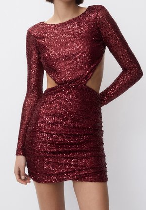 Femme portant une mini-robe bordeaux à sequins avec manches longues, découpes latérales et une silhouette ajustée.