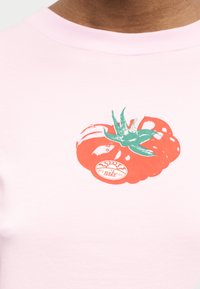 Κοντινή λήψη ροζ πουκάμισου με κόκκινο γραφικό ντομάτας και μικρό κυκλικό αυτοκόλλητο Nike στην επιφάνεια της ντομάτας.