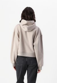 Sudadera corta beige con una gran capucha; cuenta con una textura suave y un corte relajado. Combinada con jeans de mezclilla oscuros, enfocándose en la vista posterior.