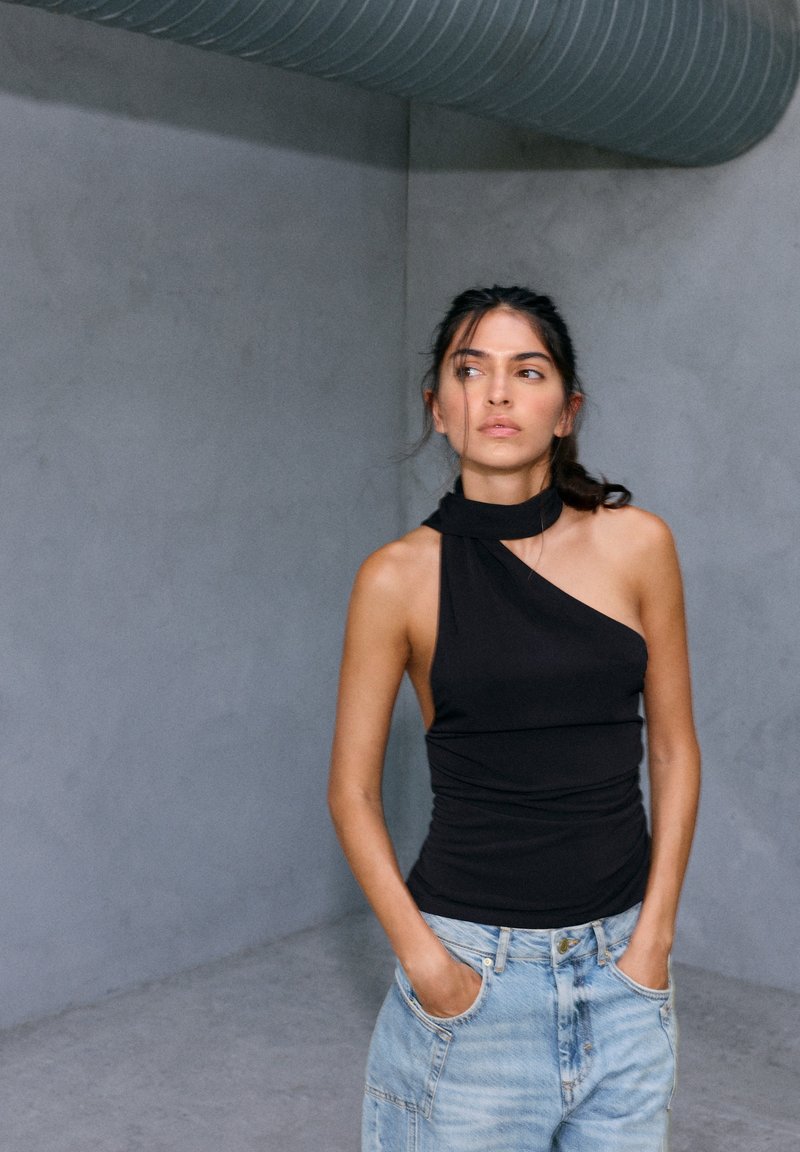 Femme aux cheveux foncés portant un top noir asymétrique et un jean bleu clair, debout les mains dans les poches, contre un mur gris avec un tuyau au plafond.