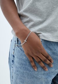 Stříbrný řetízek na zápěstí s přívěskem kříže, který má lesklý povrch. Ruka spočívá na modrých džínech a má na sobě světle šedé tričko s krátkým rukávem.