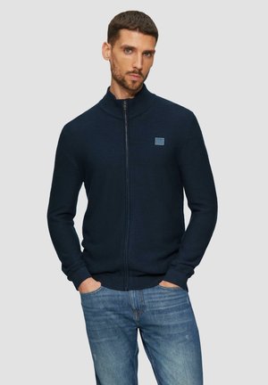 Cardigan zippé bleu marine en tissu tricoté texturé avec un col haut, arborant un petit patch logo carré sur la poitrine gauche.