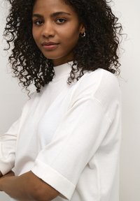 White oversized sweatshirt with short sleeves, featuring a high neck, soft texture, and minimal design. Model has curly hair.