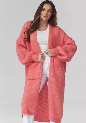 Kobieta ubrana w długi, oversize różowy sweter kardigan z dużymi kieszeniami, nałożony na biały top na ramiączkach oraz wysokowalne białe spodnie, stojąca na jednolitym tle.