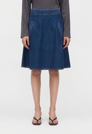 Persona con una falda vaquera azul hasta la rodilla con bolsillos delanteros, una camiseta de manga larga de color gris oscuro y sandalias negras de dedo.