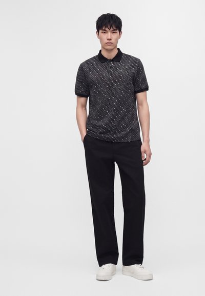 Homme portant un polo noir à manches courtes à motifs, pantalon noir, baskets blanches, debout avec une main dans la poche.