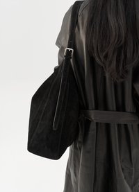 Persona con lunghi capelli scuri, che indossa un cappotto di pelle nera con cintura e porta una grande borsa a spalla in camoscio nero.