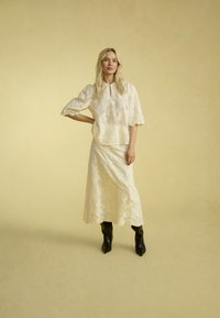 Top in pizzo crema con maniche a sbuffo e dettaglio a chiave, abbinato a una gonna in pizzo coordinata. Indossato con stivaletti neri a punta.