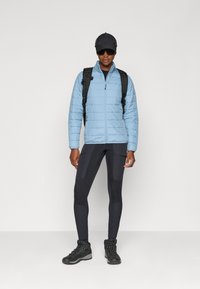 Giacca isolante blu chiaro con chiusura a zip, leggins neri e robusti stivali da escursionismo neri. Inclusi zaino con cinghia regolabile e cappellino.