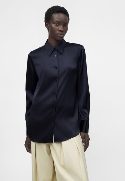 Άτομο με σκουρόχρωμο δέρμα που φορά μπλε σκούρο πουκάμισο με κουμπιά και μακριά μανίκια και φαρδιά κρεμ χρώματος παντελόνια σε απλό φόντο.