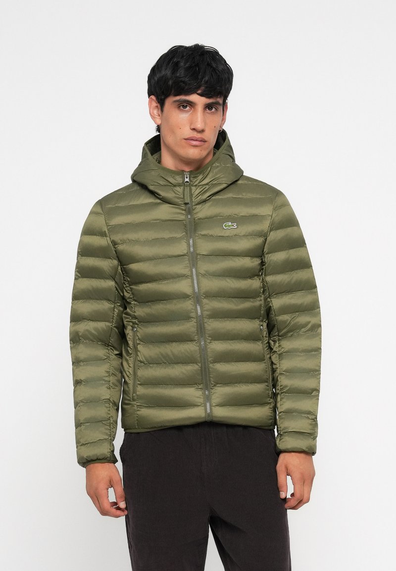 Jachetă puffer verde măslin cu glugă, având matlasare orizontală, fermoar frontal și două buzunare laterale. Sigla Lacoste pe piept.